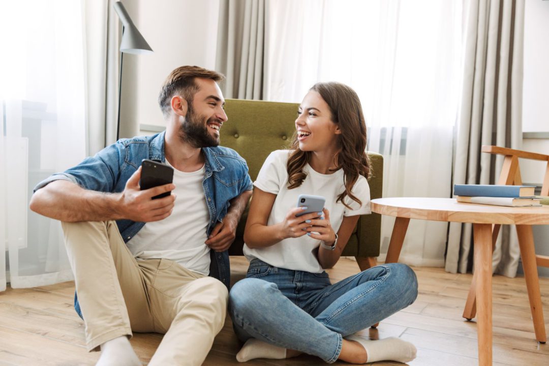 two young adults sitting in their living room and looking at each other holding smartphones in their hands