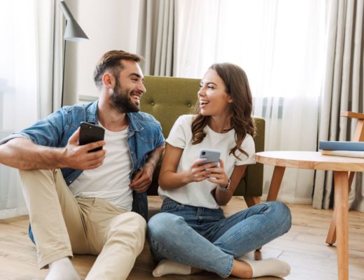 two young adults sitting in their living room and looking at each other holding smartphones in their hands