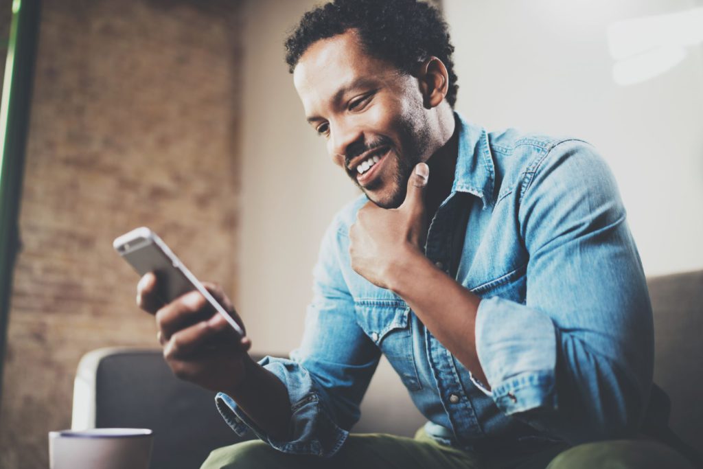 man sitting on a sofa in his home looking at his phone and smiling