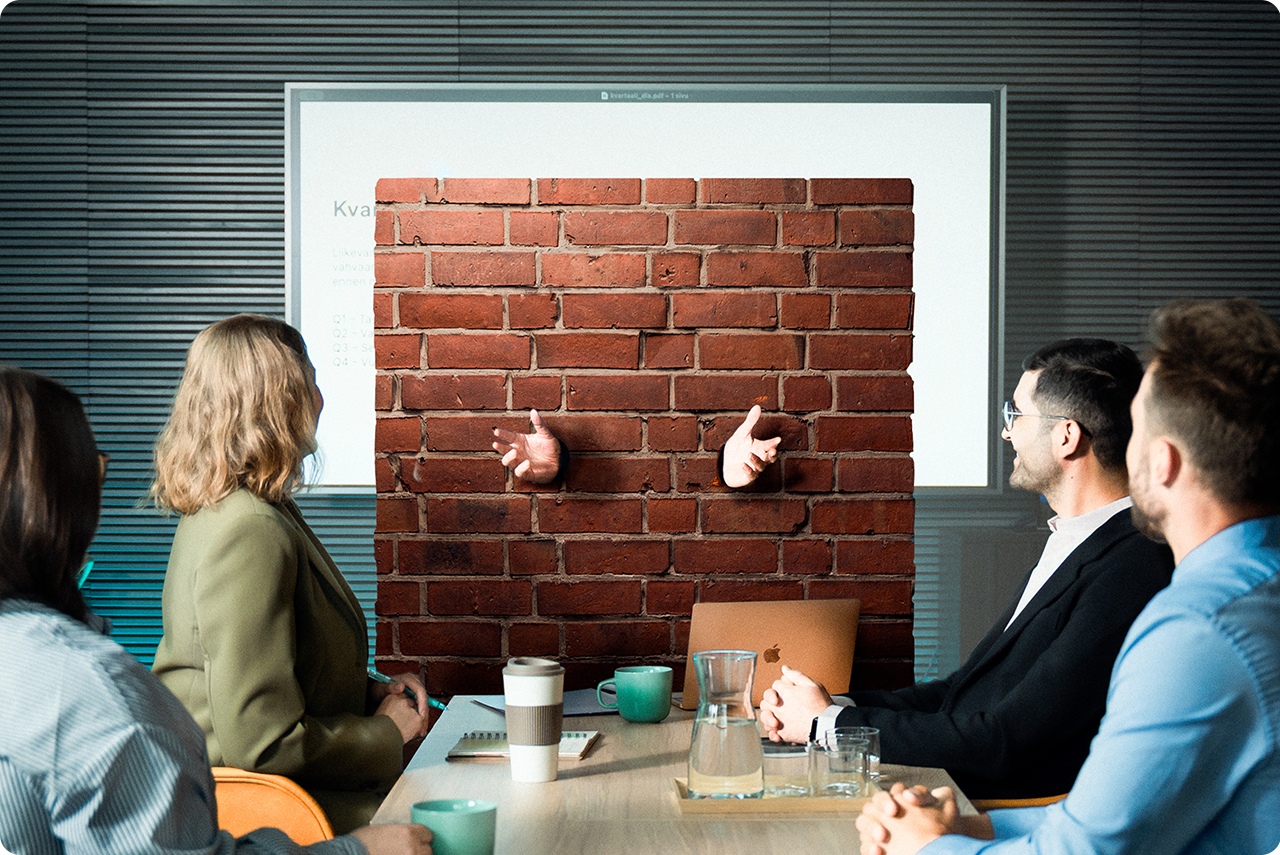 Palaveritilanne viiden ihmisen kesken. Yksi ihmisistä on tiilistä tehdyn "kielimuurin" sisällä.